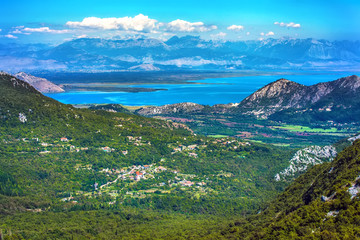 Naklejka na ściany i meble Aerial view of the coastal zone in front of the lake between the big mountains with a green forest, road and villages in the valley under the beautiful sky. Skadar Lake, Podgorica region, Montenegro.