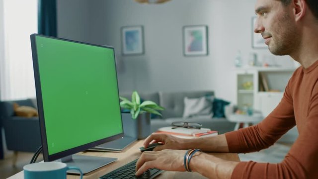 Handsome Man Sitting at His Desk at Home Uses Personal Computer with Mock-up Chroma Key Screen.