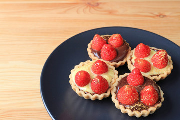 Mini chocolate tarts topped with fresh strawberries and edible gold powder served on black plate on wooden table with free space  
