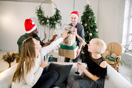 Picture showing group of friends with Christmas presents, celebrating Christmas or New Year in cozy decorated living room. Two boys, African and Caucasian in santa hats with two pretty Caucasian girls