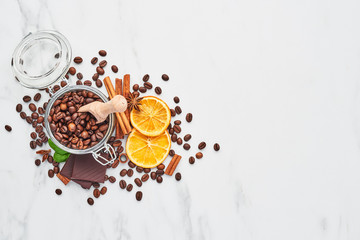 Coffee in a glass jar with beans, chocolate, dried oranges, anis and cinnamon sticks on white marble background. Concept of coffee with different spices. Top view with copy space for text.