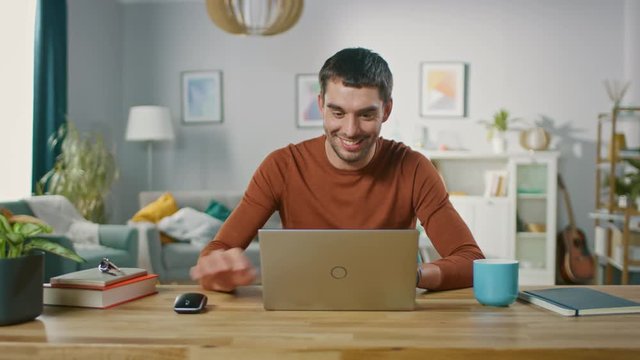 Confident Young Man Sitting at His Desk at Home, Opens and Starts Using Laptop. In the Background Living Room with Cozy Design.