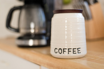 Close Up Bright Modern Stock Photo of Cute White Coffee Bean Container onto of Wooden Counter Top with Coffee Maker and Knife Set Blurred in Background of Elegant Kitchen with Natural Light