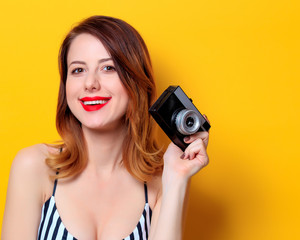 Portrait of beautiful young smiling redhead white woman in black striped dress eith retro camera on yellow background