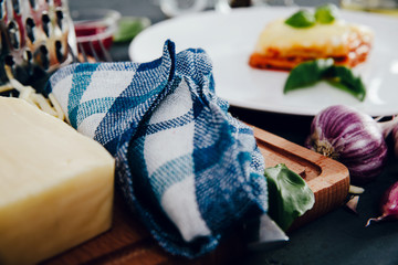 Cutlery in a blue checkered napkin on a stone counter, in the background a piece of lasagne on a white plate. Preparation of lasagna. Adding spices to food, enhancing taste, spicy flavors.