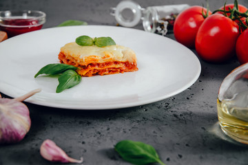 Lasagne on a white plate. On the stone table lies lasagna, spices and food ingredients. The concept of eating and preparing lasagna in tomato sauce.