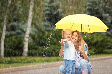 Portrait of happy mother and daughter with yellow umbrella in park on rainy day. Space for text