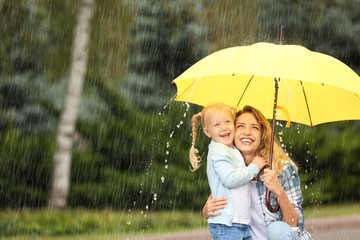 Portrait of happy mother and daughter with yellow umbrella in park on rainy day. Space for text