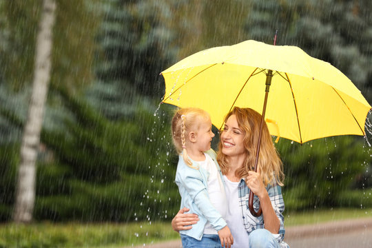 Portrait of happy mother and daughter with yellow umbrella in park on rainy day. Space for text