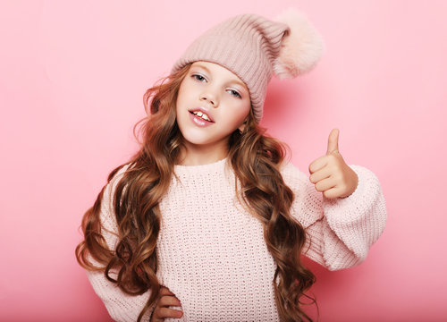 People, Childhood And Fashion Concept: Little Girl Child Wearing Pink Winter Hat And Sweater Over Pink Background