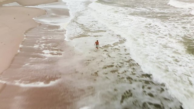 Aerial Fly Over Of A Beach Bro Holding His Surfboard Walking Through The Breakwater At The Beach.