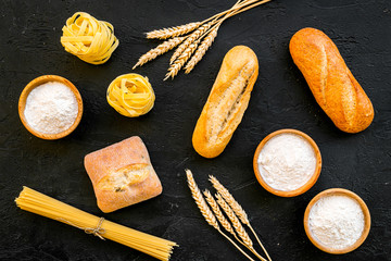 Homemade fresh bread and pasta near flour in bowl and wheat ears on black background top view