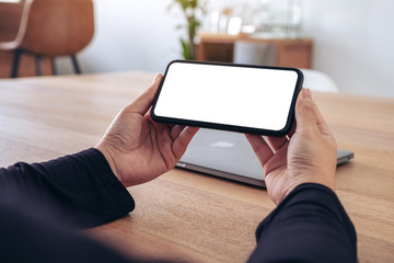 Mockup image of hands holding black mobile phone with blank white screen horizontally for watching with laptop on table in office