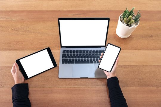 Top view mockup image of a hands holding mobile phone , black tablet and laptop with blank white screen on wooden table in office