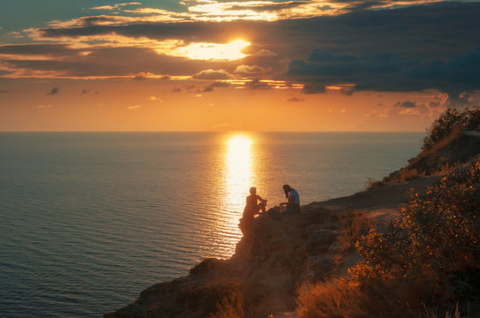 Пара туристов сидят на краю обрыва скалистого побережья Черного моря на закате солнца. Мыс Фиолент, Крымский полуостров