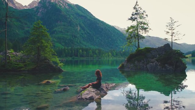 In this Cinemagraph a young brunette woman is sitting on a rock at the scenic lake Hintersee in Bavaria, Germany with the water moving gently.