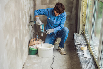 Young male painter kneads putty with water in a bucket using a hand-held mixer for building mixes