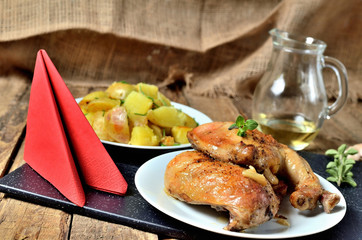 Golden grilled chicken legs on a plate and roasted potatoes, rosemary, jug with oil, oregano, salt, pepper, garlic and red napkin on a wooden table