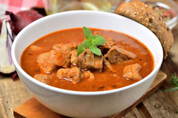 Close-up of pork goulash in a bowl, oregano pepper, onion, red checkered tablecloth in the background