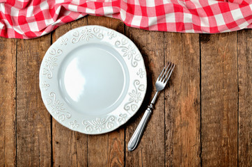 Decorative ceramic plate, fork and red checkered tablecloth on top side on old vintage wooden table background - top view