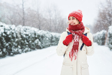 Cheerful smiling woman in warm clothes, red knitted cap, scarf and mittens walking on the snowy street under falling snowflakes after blizzard in city. Happy woman playing with the snow