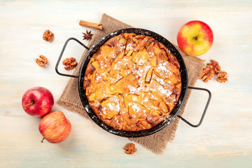 A photo of an apple pie in a pan, shot from the top on a rustic wooden background with apples, cinnamon, walnuts and copy space