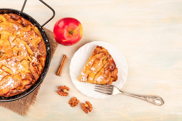A photo of an apple pie in a pan and a slice of it on a plate, shot from the top on a rustic wooden background with an apple, a cinnamon stick, walnuts, and a place for text