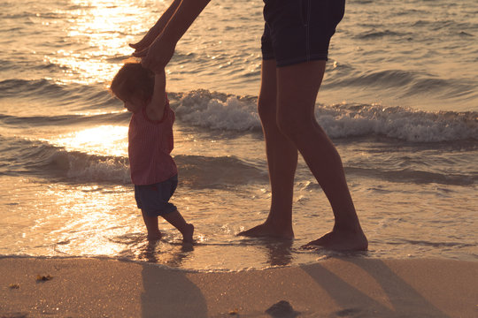 Father helping his baby with the first steps, teaching baby to walk concept, outdoor candid photo on the beach, spending a day at the beach, healthy family lifestyle.