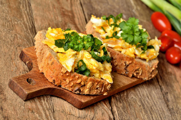 Close-up of wholegrain slices of bread with scrambled eggs, fresh herbs, spring onions and tomatoes in background