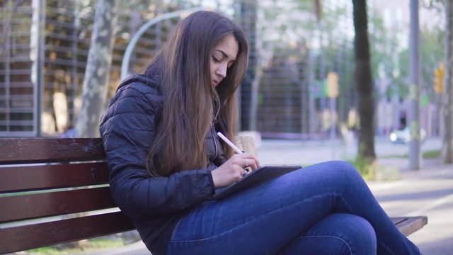 4k shot of a woman drawing on digital tablet with stylus pencil. Morern designer and paint freelance artist. Outdoors on the bench