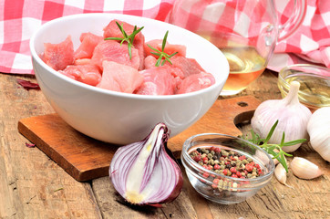 Raw pork meal diced in a bowl, garlic, half of onion, pepper, jug with oil and checkered red tablecloth in the background