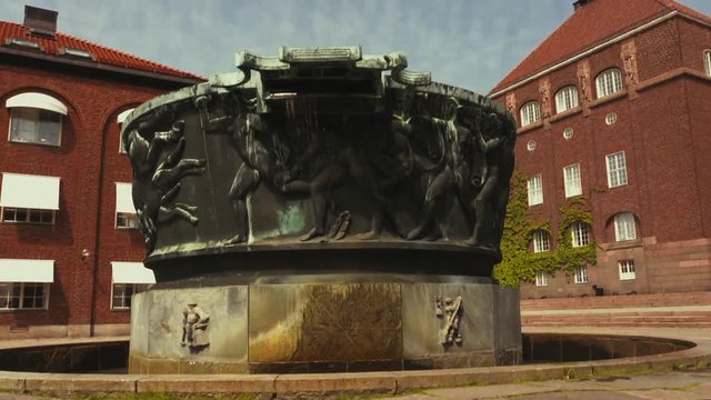 A large decorative fountain in the center of a campus courtyard. Ornate sculpture around the outside of a water feature near brick buildings. Wind blows the water spilling out the side. Closeup view.