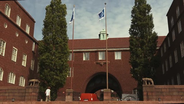 A red van drives through an iron gate at the entrance to a university campus as a pedestrian walks past. Dark red brick building with flags of KTH Royal Institute of Technology flying from poles.