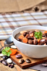 Bowl of bean soup with large beans on cutting board, carrots, parsley, marjoram, spoon and ladle, towel in background - vertical photo
