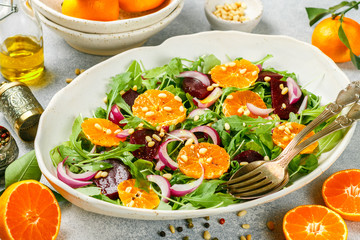 Delicatessen colorful salad of baked beets, arugula, tangerines and red onions with spices and pine nuts in a white dish. Olive oil and ingredients on a grey surface