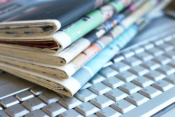 Newspapers and laptop. Pile of daily papers with news on the computer. Pages with headlines, articles folded and stacked on keypad of electronic device. Modern gadget and old journals, focus on paper  - obrazy, fototapety, plakaty