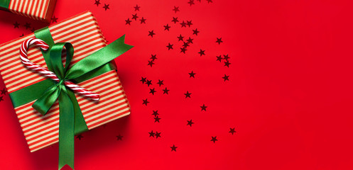 Obraz premium Gift boxes with green ribbon, Candy Cane, glitter confetti form of stars on red background top view flat lay. Holiday concept, new year or Christmas gift box, presents Xmas holiday