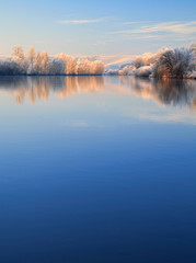 Naklejka na ściany i meble Winter Landscape Reflecting in River at Sunrise, Trees covered by hoarfrost and snow, blue sky