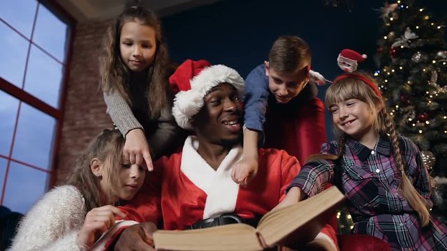 Portrait shot of african boy in the santa claus costume reading book with kids.
