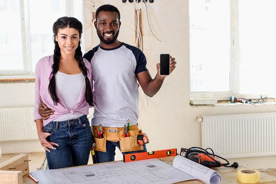 african american couple showing smartphone with blank screen during renovation at new home