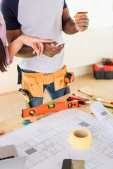 partial view of woman pointing by finger on smartphone while her boyfriend standing near with credit card near table with blueprint during renovation of home