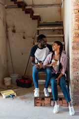 Fototapeta na wymiar smiling african american couple resting with disposable cups of coffee during renovation at new home