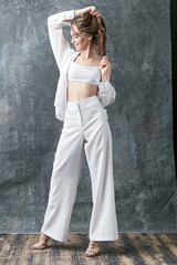 Fototapeta premium Smiling brunette female model posing in white shirt, top and pants. Studio shot, grey background