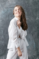 Smiling brunette female model posing in white shirt, top and pants. Studio shot, grey background