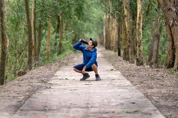 Asian runner in sport suit are running with happiness action drinking the water from sport bottle with relax action in deep forest in evening time, lifestyle and leisure concept