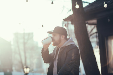 portrait of handsome man with beard in hat in leather jacket with holding cup of hot coffee. 