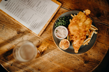 Traditional english fish and chips plate on a wooden table, with a pint and a menu.