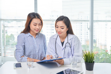 Female doctor talks to female patient in hospital office while writing on the patients health...