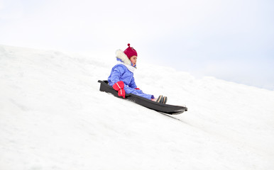 Fototapeta na wymiar childhood, sledging and season concept - happy little girl sliding on sled down snow hill outdoors in winter