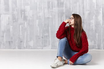 Isolated shot of beautiful 16 year old female in knitted cozy sweater, eyeglasses, jeans and sneakers sitting on floor and looking away with pensive facial expression, thinking about teenage problems